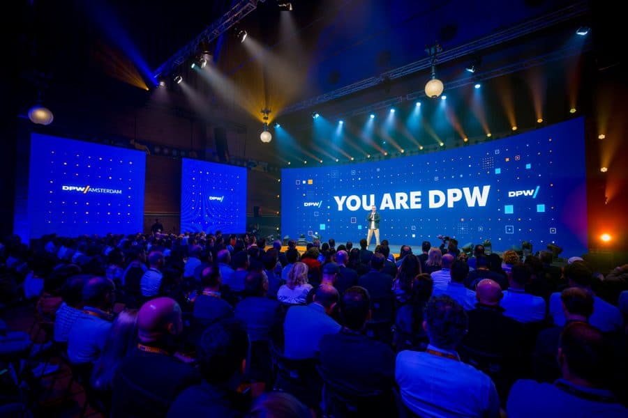 Speaker on the DPW Amsterdam main stage before a packed hall, with "YOU ARE DPW" on the big screen.